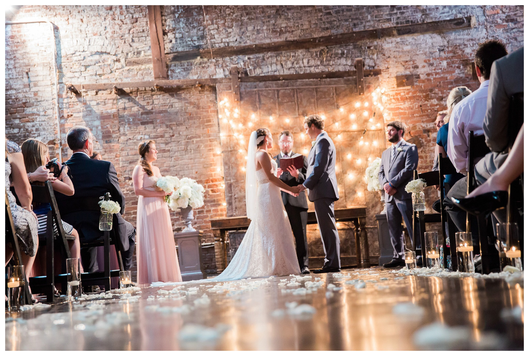 A perfect Scene - The Vows shot from the shimmering floor toward the industrial brick wall behind the couple and the pastor,  Venue and Catering – King plow Event Gallery and Bold American Events Decor and flowers – Stylish Stems Music and Band – Seven Sharp Nine Transportation – Georgia Trolley Photographer One Life Photography