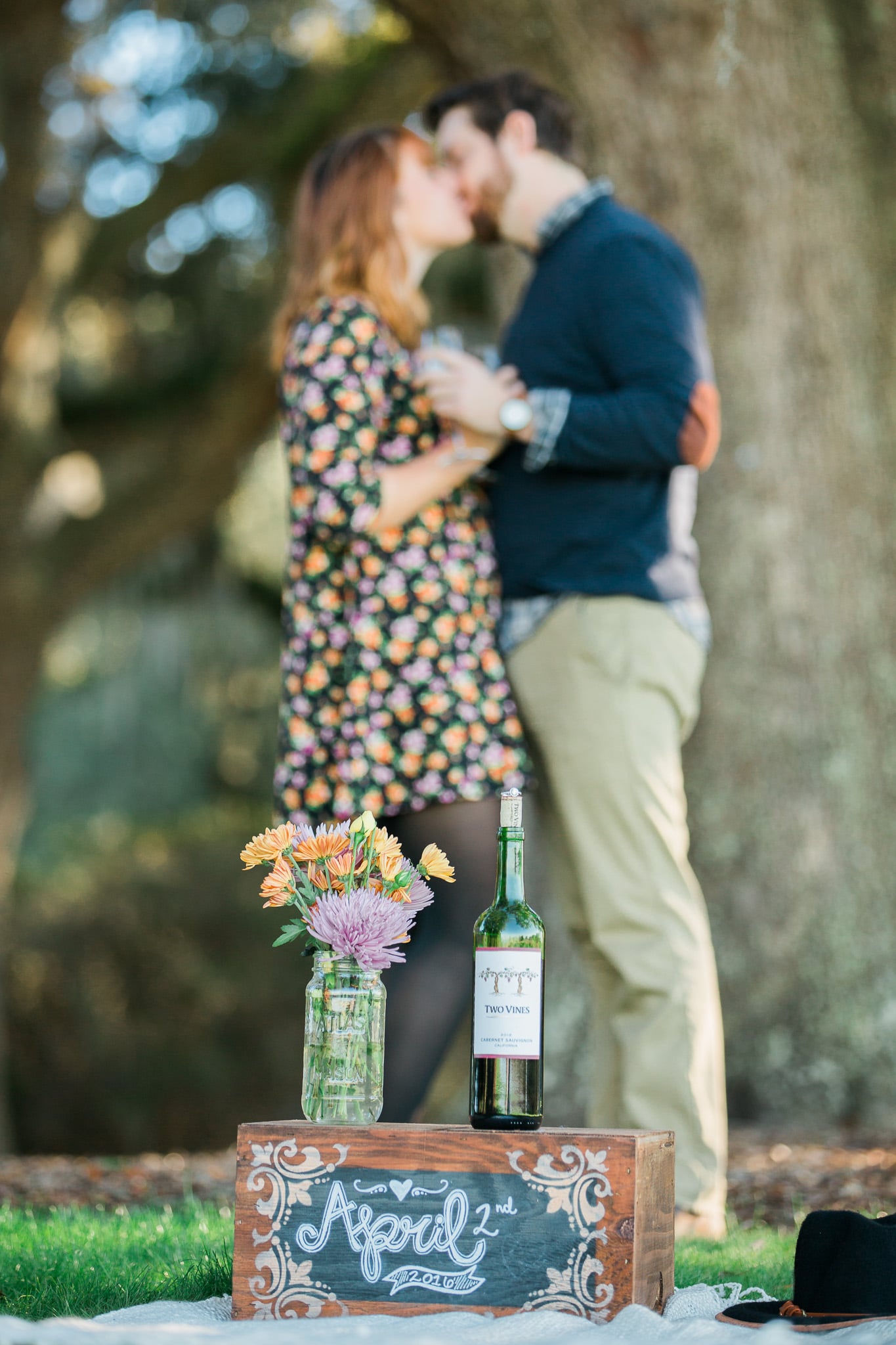 Brian and Kelsey out of focus in the background kissing with the wedding notice bouquet and wine in the foreground crystal clear, Caledonia Golf and Fish Club, Myrtle Beach Engagement Session 9, www.onelifephoto.net